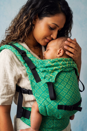 Close three-quarter profile of a mother resting her cheek against her sleeping baby nestled in the green Almitra Tattva carrier