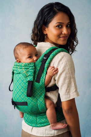 Over-the-shoulder view of a woman carrying her baby on her back in the green geometric woven Almitra Tattva baby carrier.