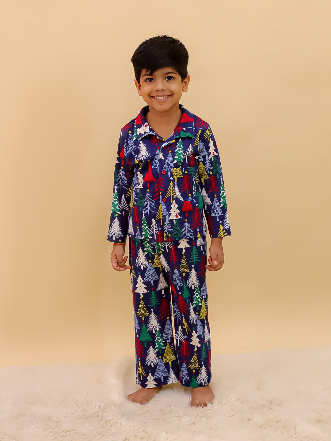 Little boy wearing The Mom Store Silent Night Christmas Kids Winter Pajama Set — a navy blue pajama set with colorful Christmas tree prints, standing barefoot on a fluffy white rug and smiling.