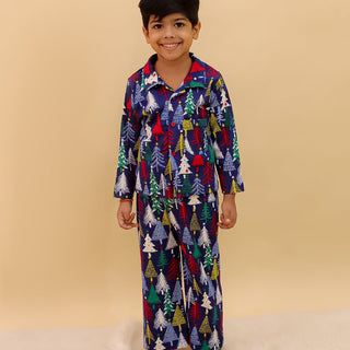 Little boy wearing The Mom Store Silent Night Christmas Kids Winter Pajama Set — a navy blue pajama set with colorful Christmas tree prints, standing barefoot on a fluffy white rug and smiling.