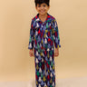 Little boy wearing The Mom Store Silent Night Christmas Kids Winter Pajama Set — a navy blue pajama set with colorful Christmas tree prints, standing barefoot on a fluffy white rug and smiling.