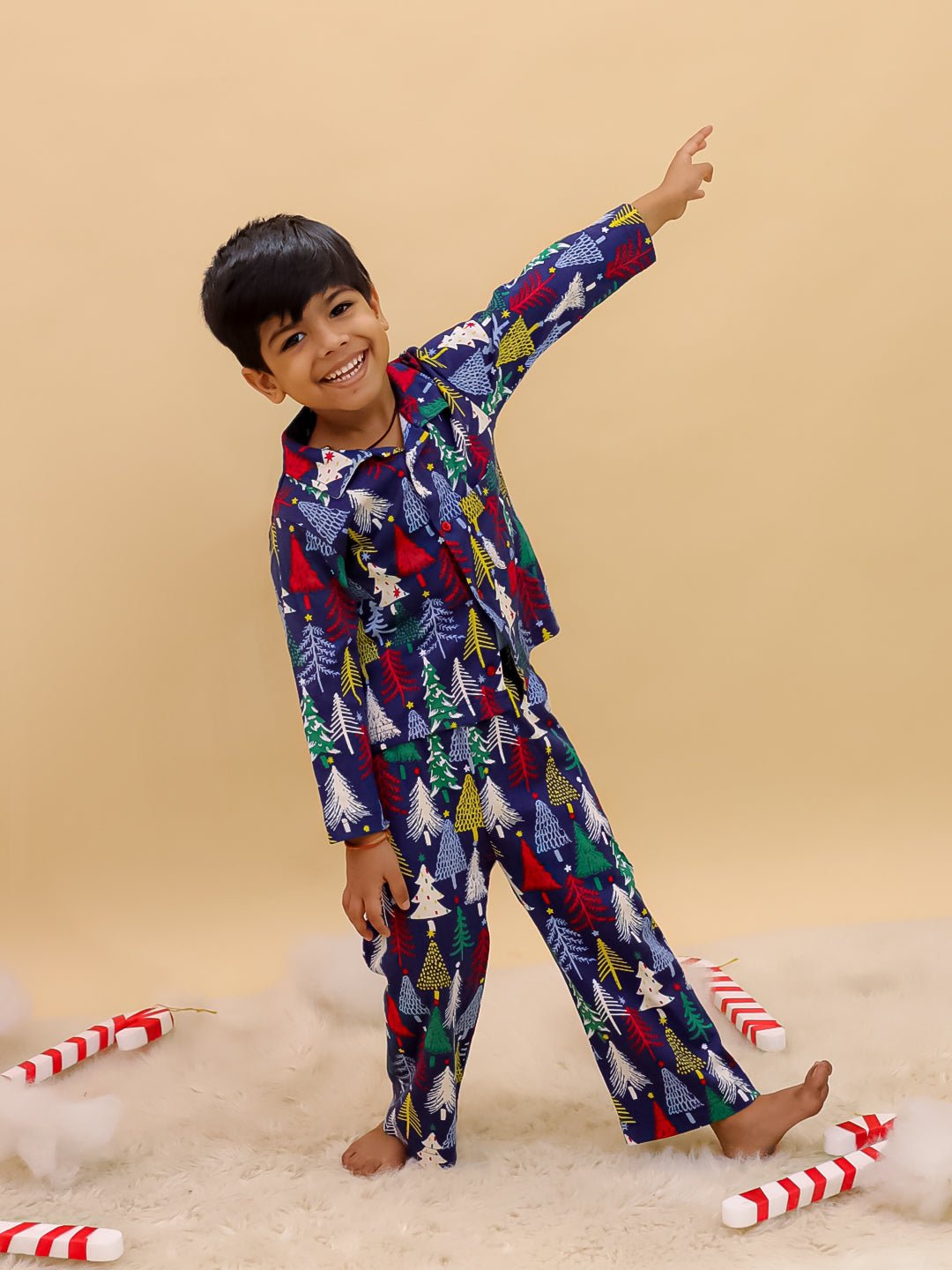 Boy in The Mom Store Silent Night Christmas Pajama Set posing joyfully with one arm stretched out, surrounded by candy cane props and cotton clouds on the floor.