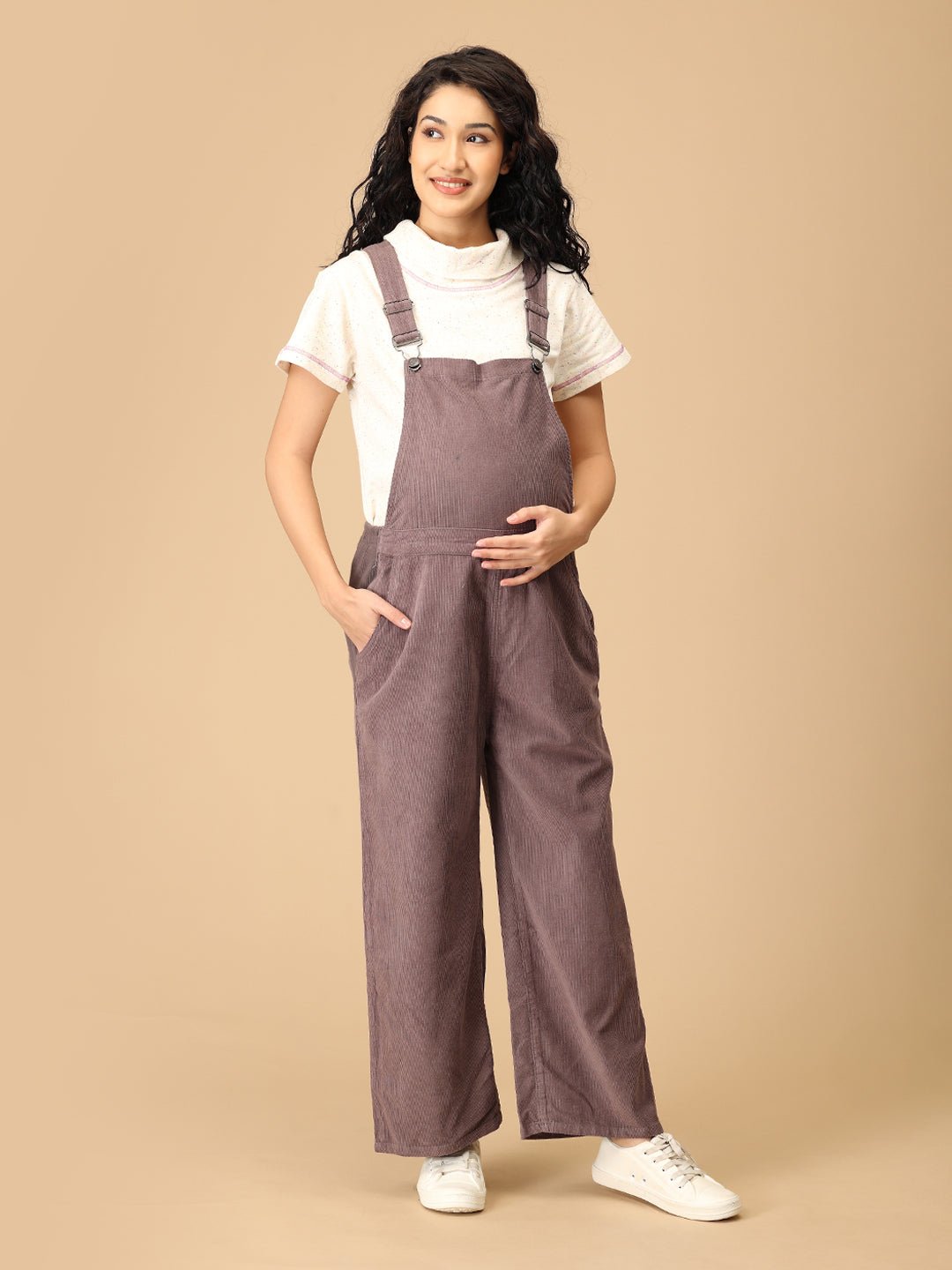 Pregnant woman posing in The Mom Store taupe corduroy maternity dungaree with adjustable straps.