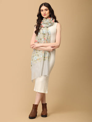 A woman wearing a white dress and brown boots, standing and posing with a floral fine wool stole with embroidery and a tasseled border.
