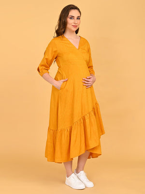 Portrait of a lady wearing Yellow a go go Maternity Wrap Dress, standing against orange background with her hands inside the pockets